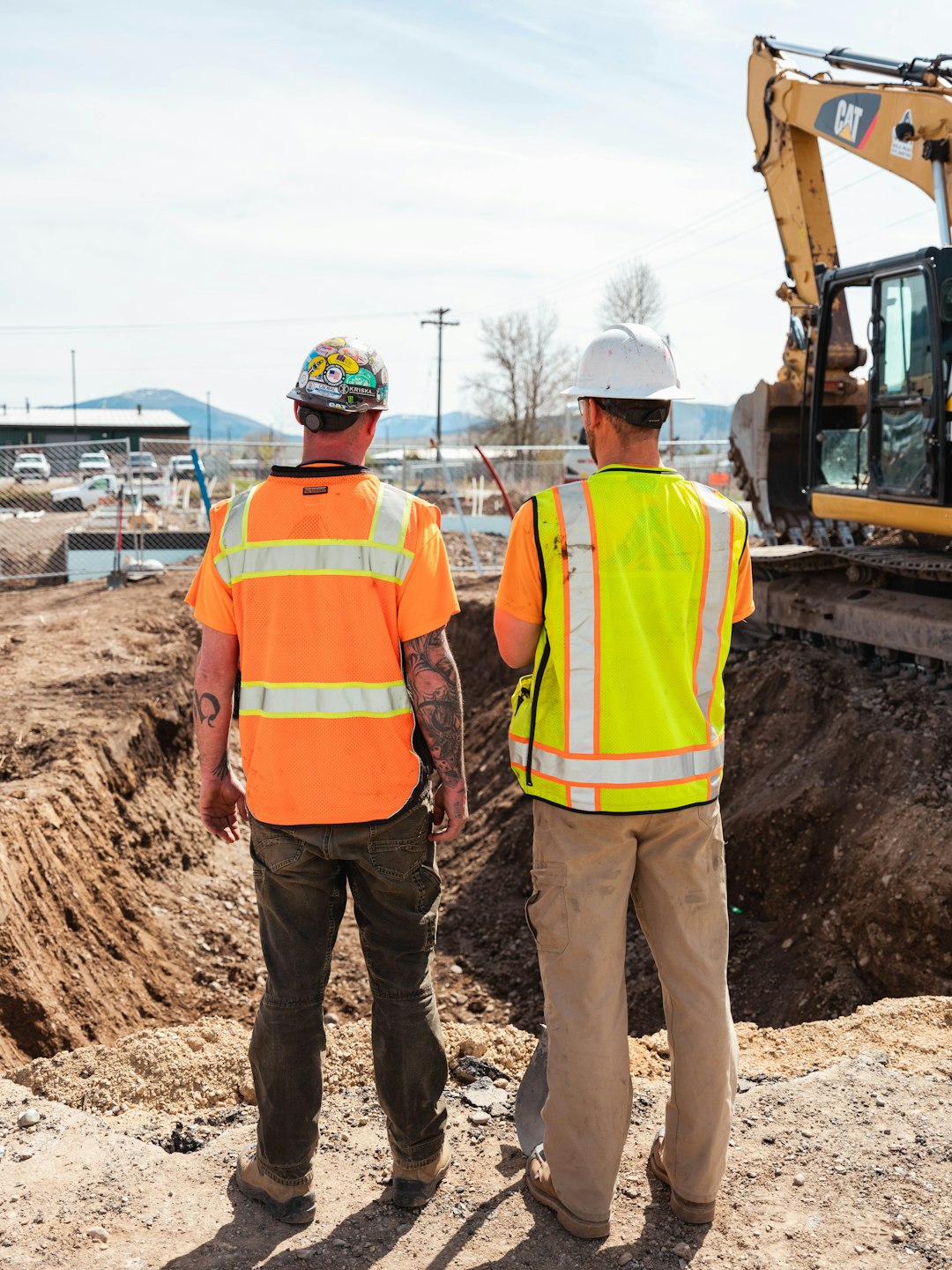 Home Two construction workers in high-visibility safety clothing review an in-progress excavation at a jobsite.