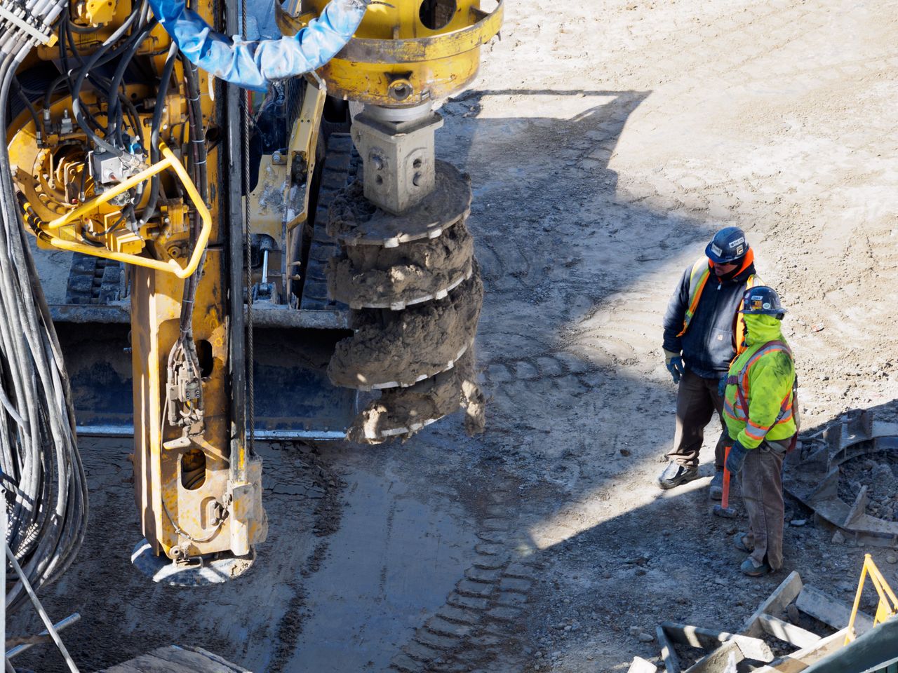 Two workers in protective gear at a construction site with heavy drilling machinery.